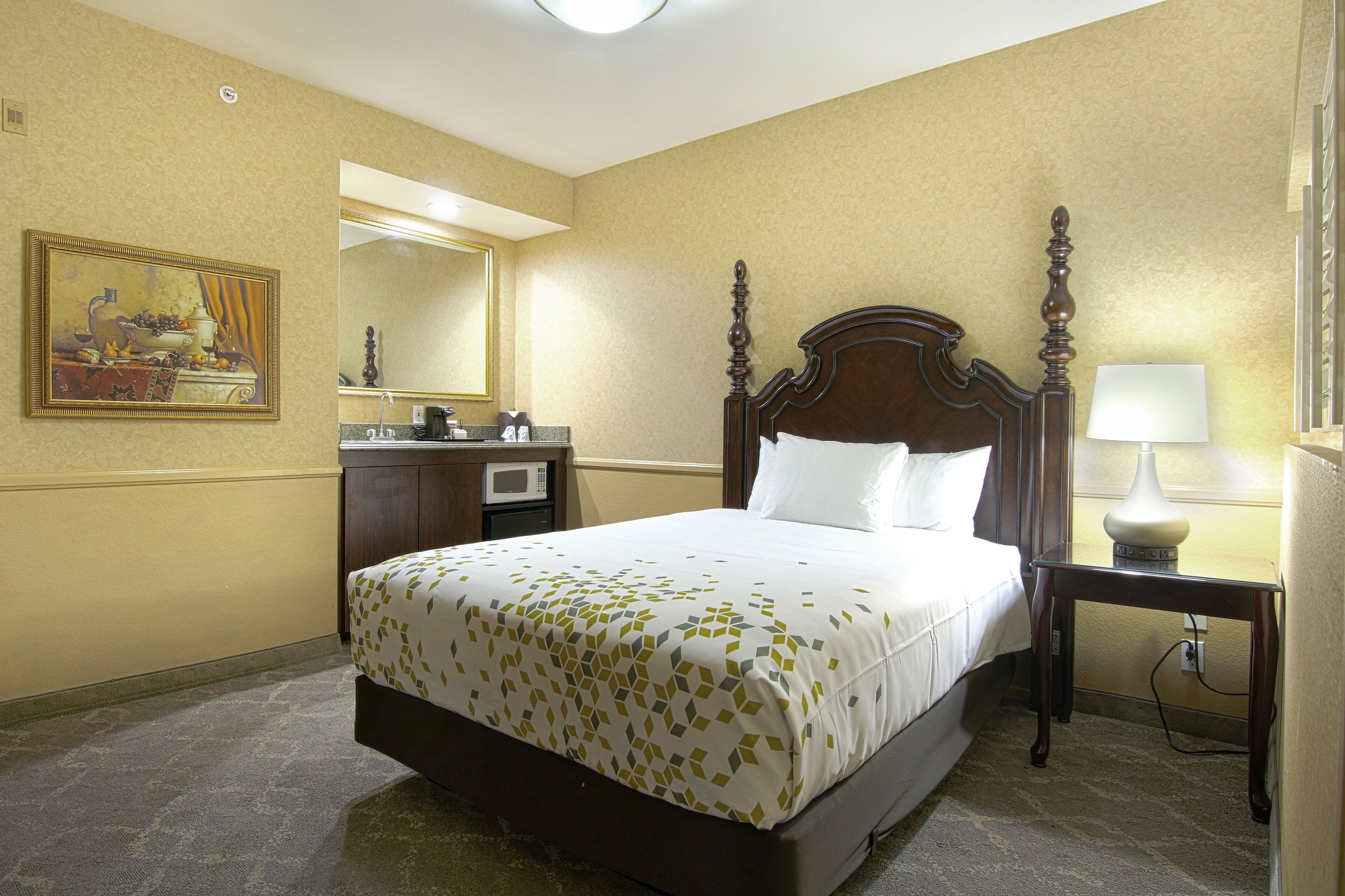Un cuarto de hotel con una cama grande, un cabecero de madera, una mesita de noche con lámpara, un aparador, un microondas y un cuadro en la pared.
