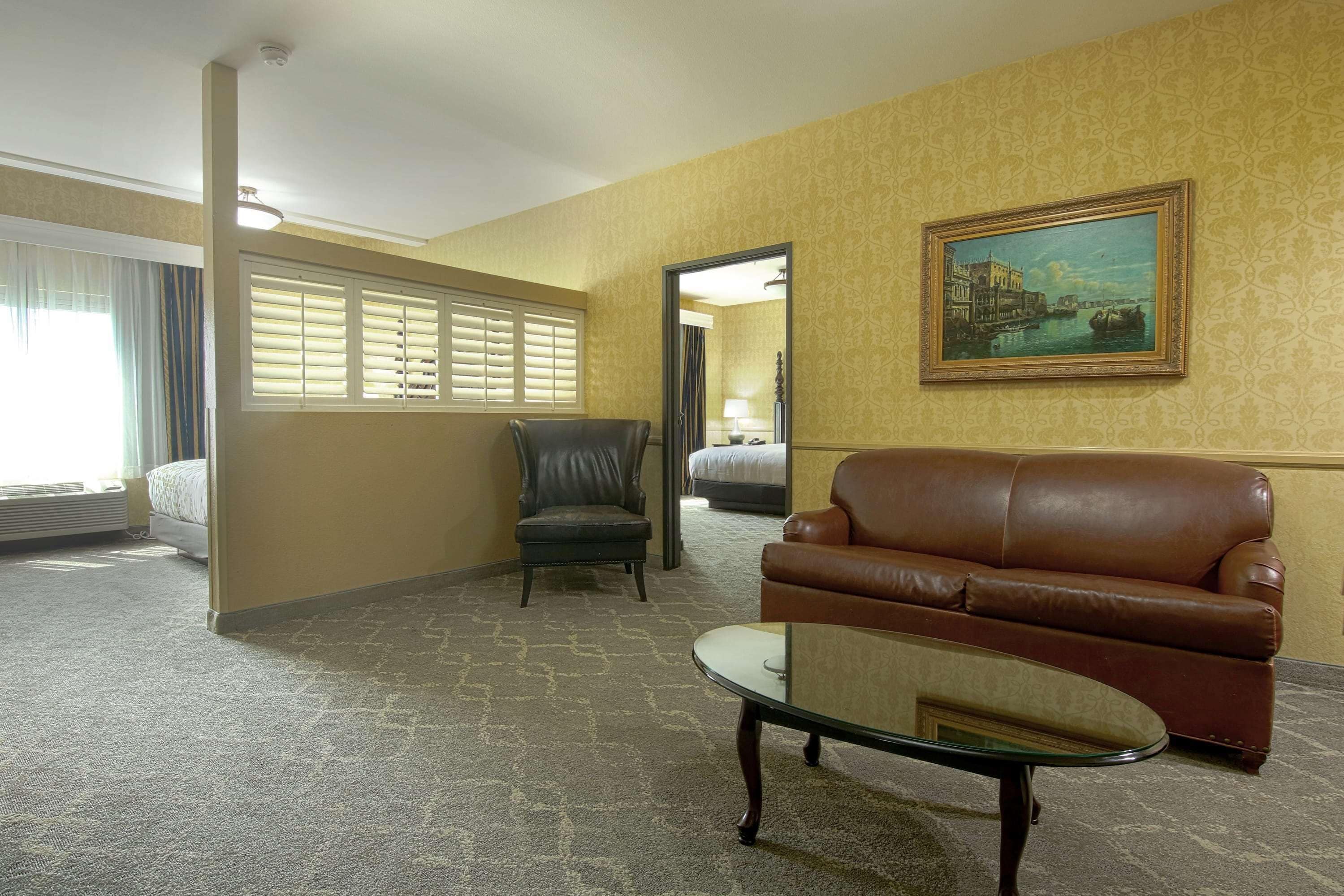 A hotel room with a brown leather sofa, a glass coffee table, a leather armchair, a bed with a white comforter, a painting on the wall, and a window with blinds.