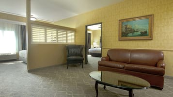 Une chambre d’hôtel avec un canapé en cuir marron, une table basse en verre, un fauteuil en cuir, un lit recouvert d’une couette blanche, un tableau au mur et une fenêtre avec des stores.