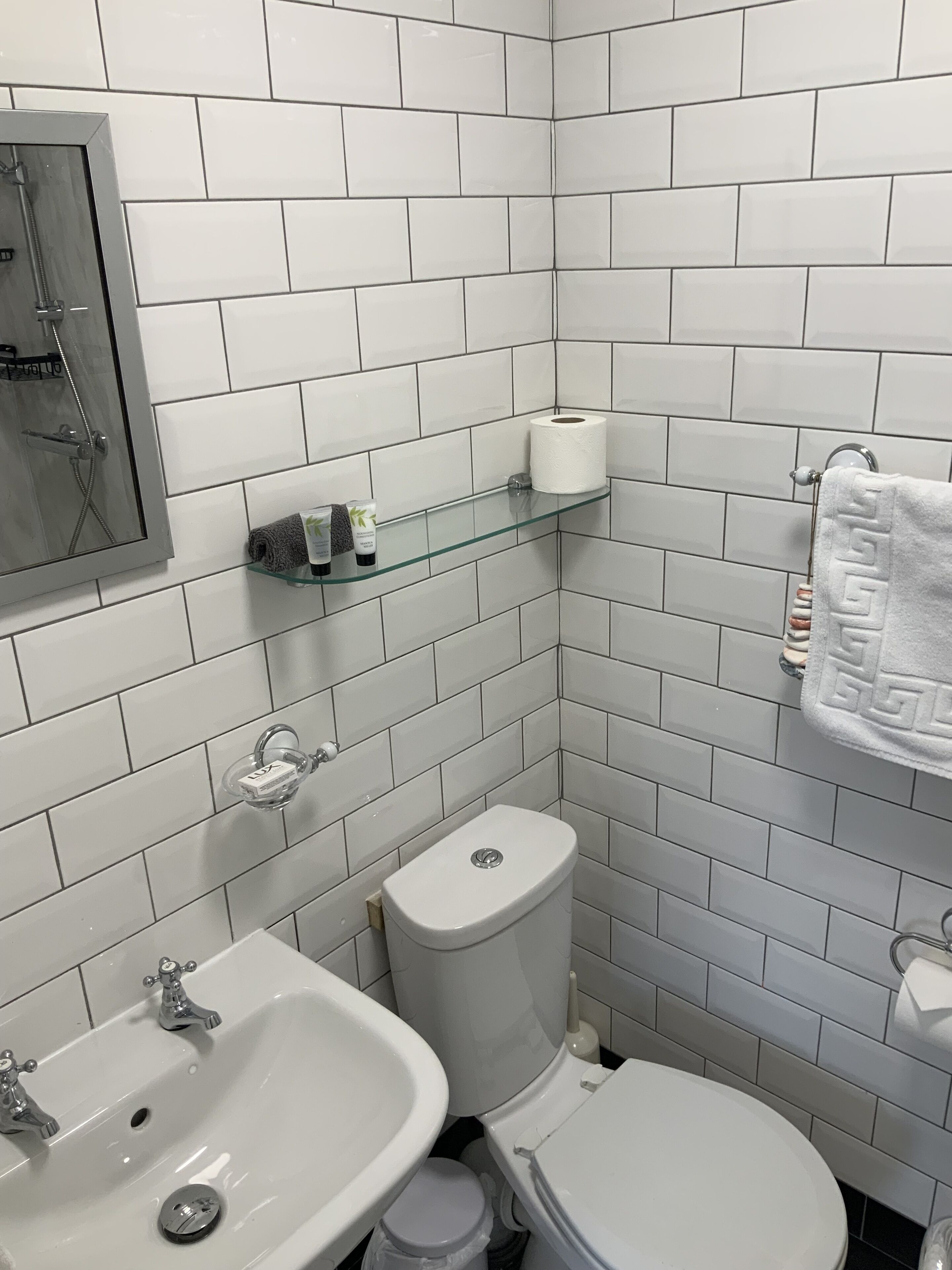 A bathroom with white tiles, a glass shelf with toiletries, a toilet, and a sink.