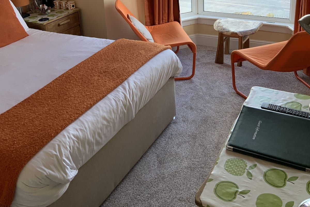Habitación de hotel con una cama, dos sillones naranja, una mesita con un ordenador portátil y vistas al mar a través de amplias ventanas.