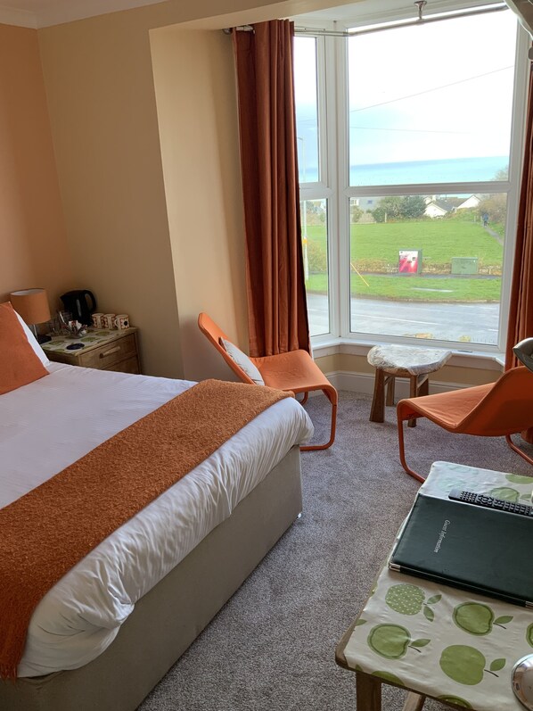 A hotel room with a bed, two orange chairs, a small table with a laptop, and a view of the sea through large windows. - Green Apple Bed and Breakfast (St Ives)