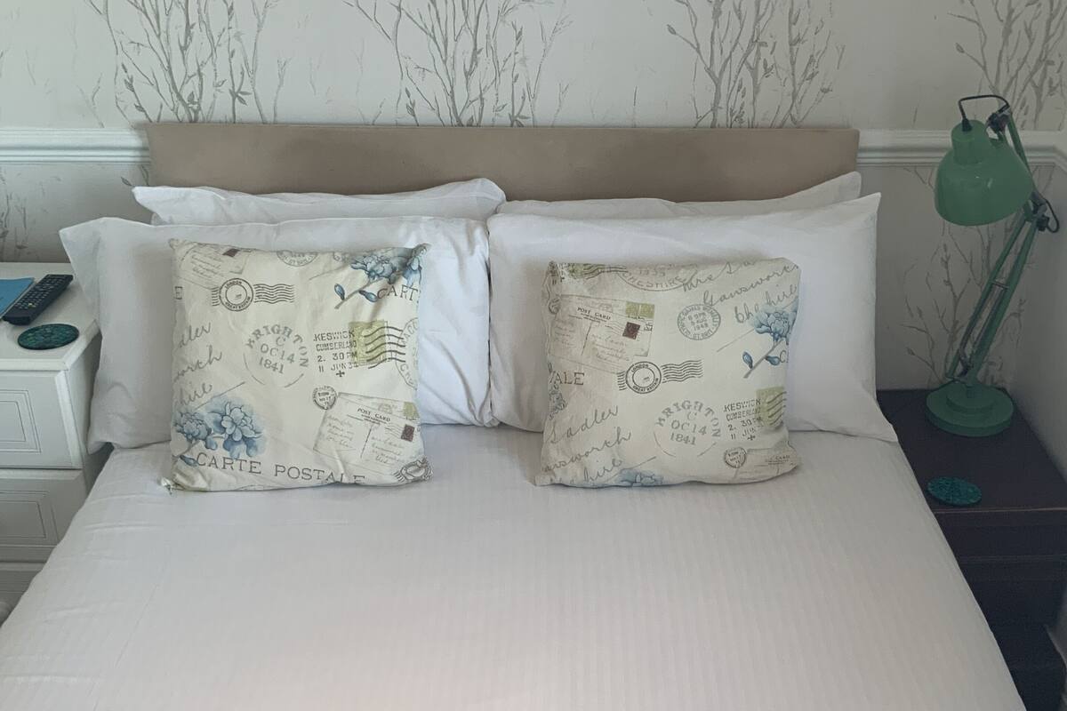 Una cama con dos almohadas, una mesita de noche con lámpara y una pared con diseño de árbol desnudo.