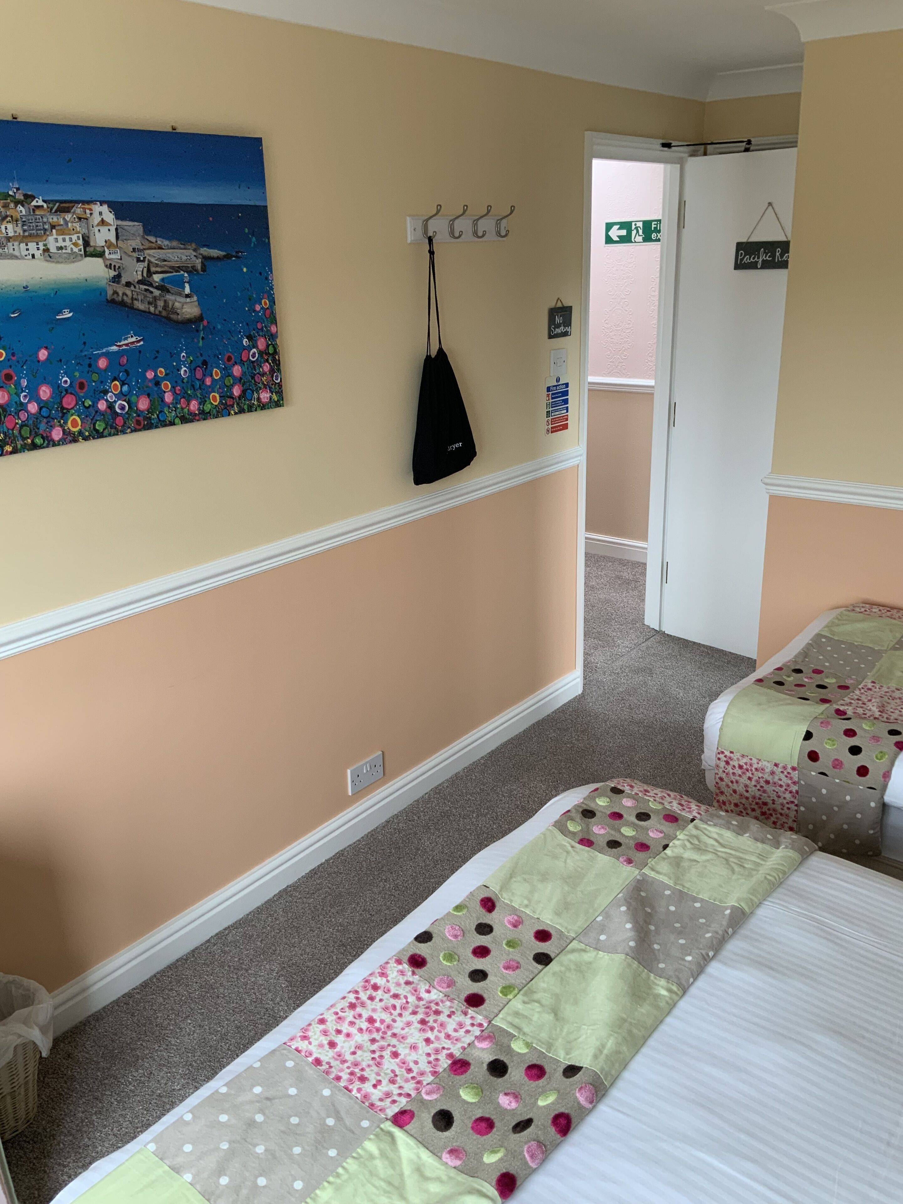 A hotel room with two beds, a painting of a coastal scene, a coat rack, and a door leading to another room.