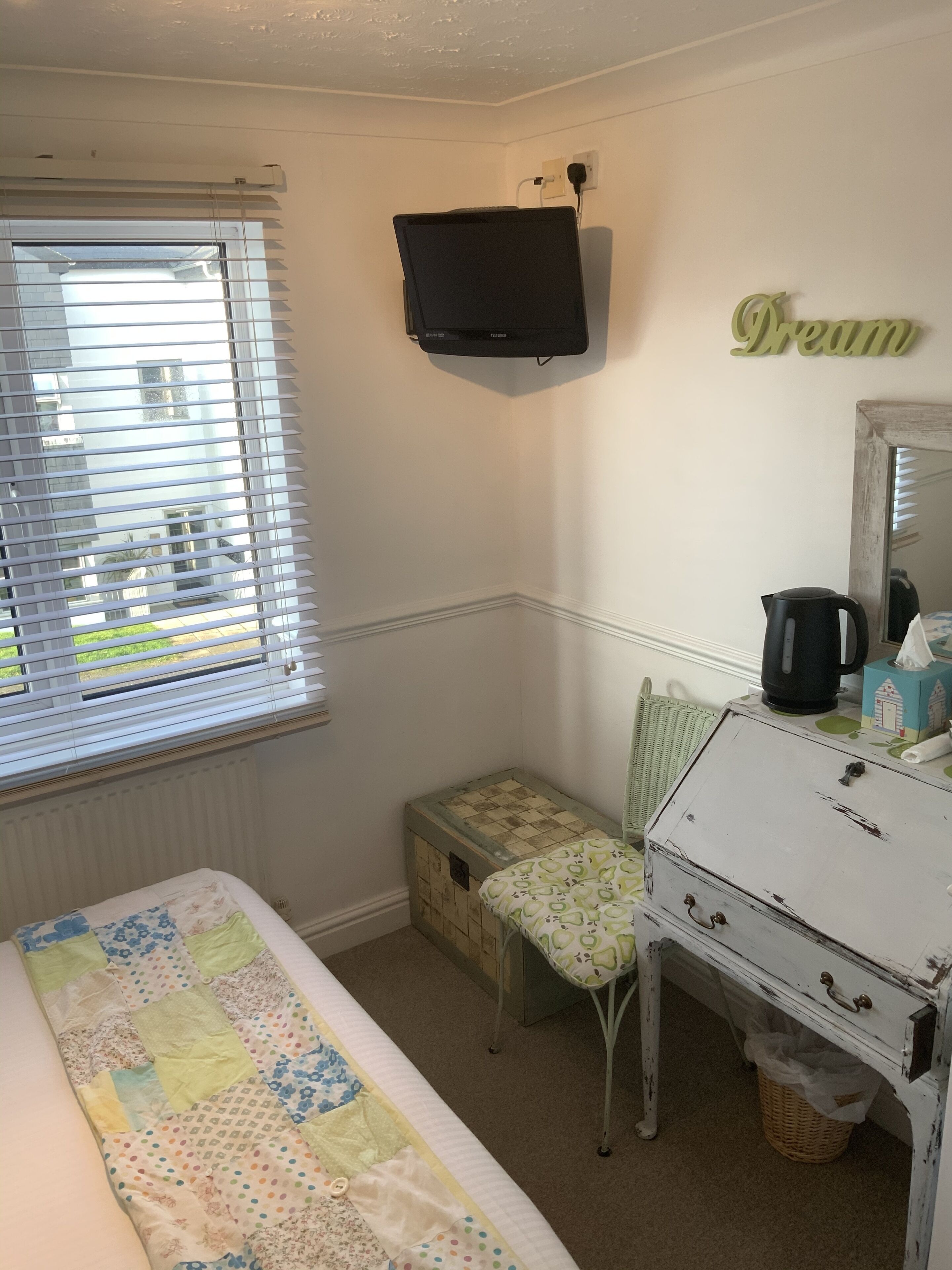 A bedroom with a bed, a dresser, a chair, a small table, a television, and a window with blinds.