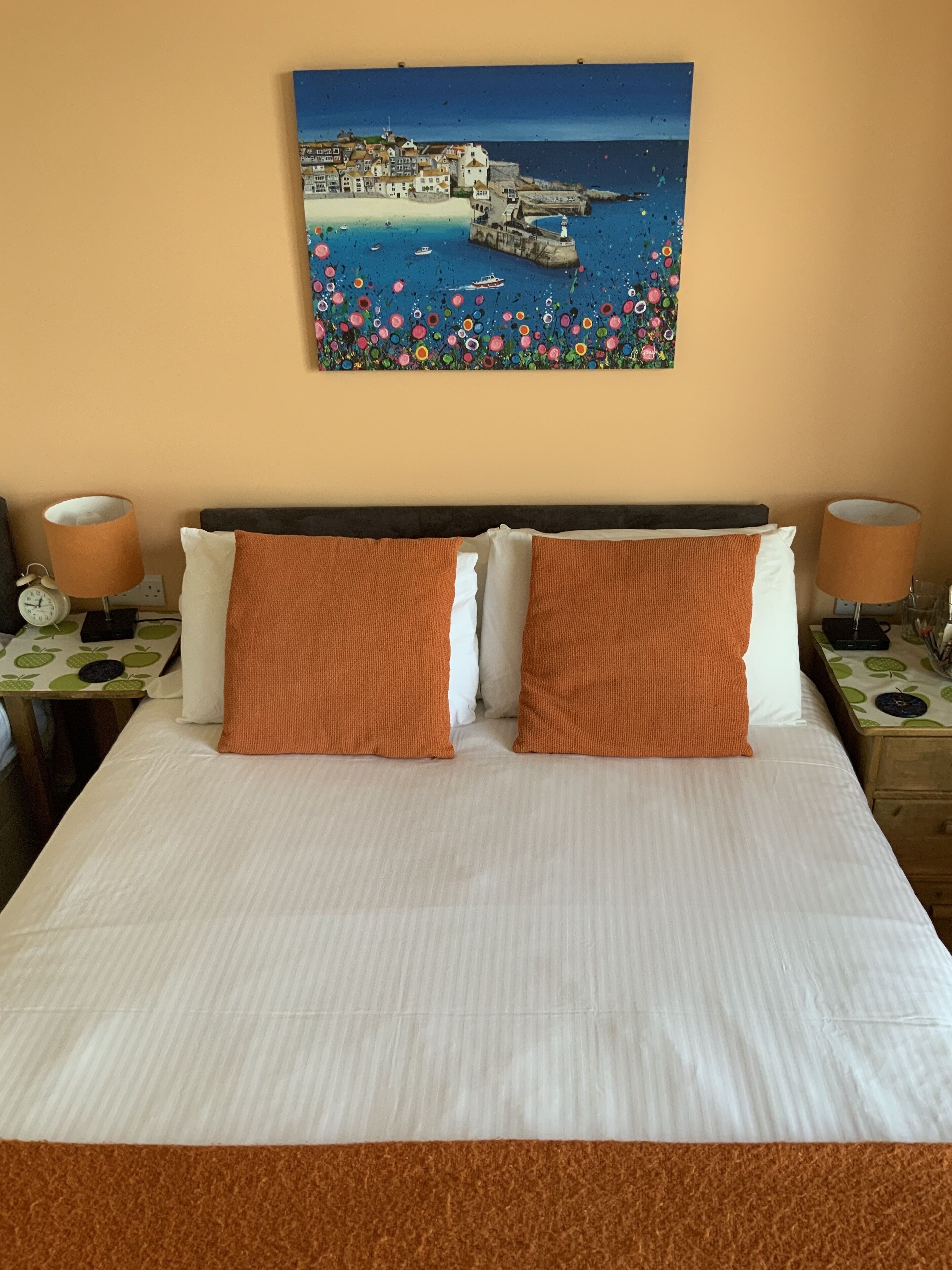 A bed with white linens, two orange pillows, and a painting of a coastal scene on the wall.