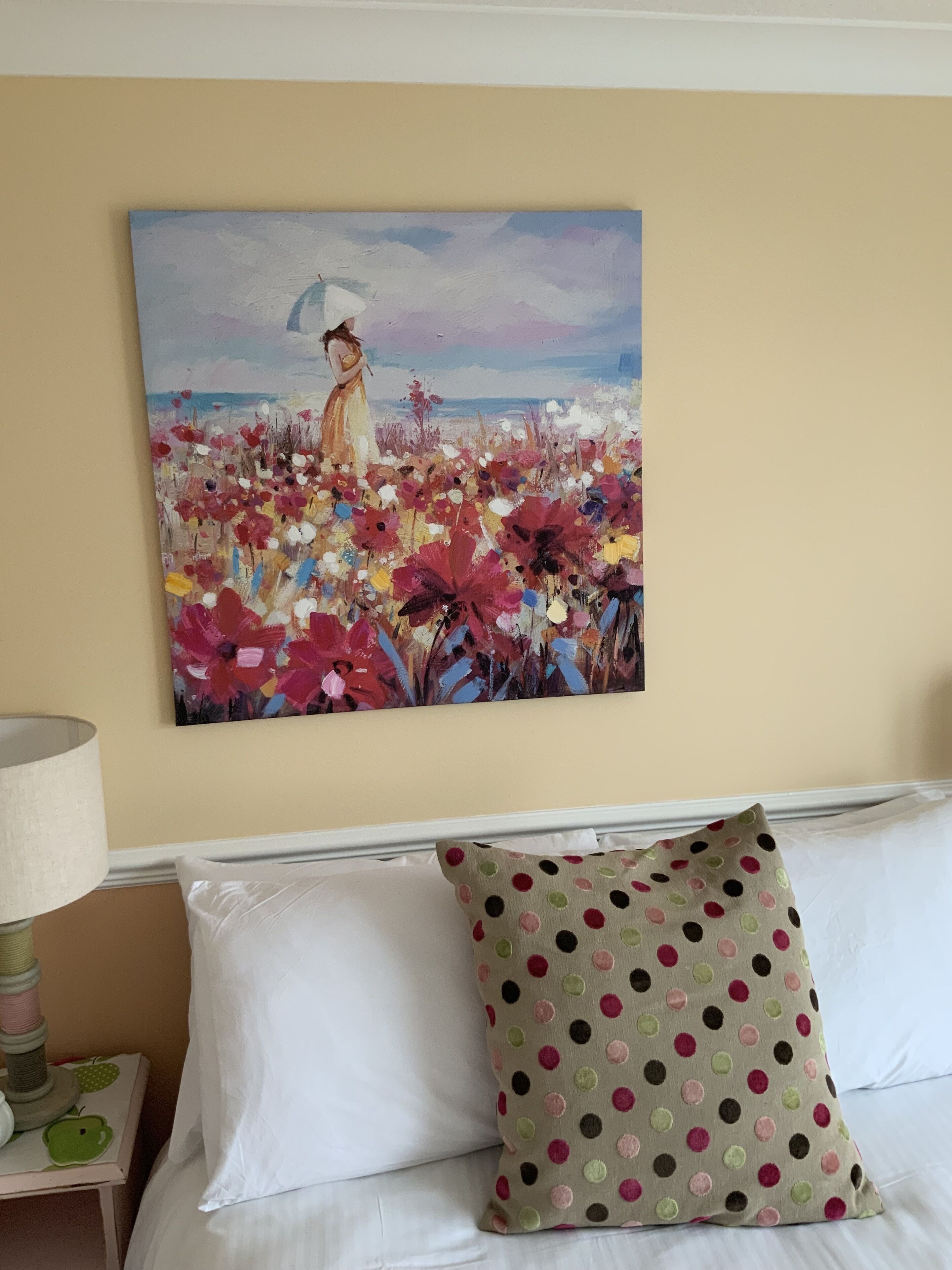 A painting of a woman in a field of flowers on a wall, a white couch with a polka dot pillow, and a lamp on a bedside table.