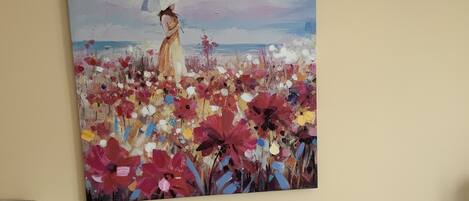 A painting of a woman in a field of flowers on a wall, a white couch with a polka dot pillow, and a lamp on a bedside table.