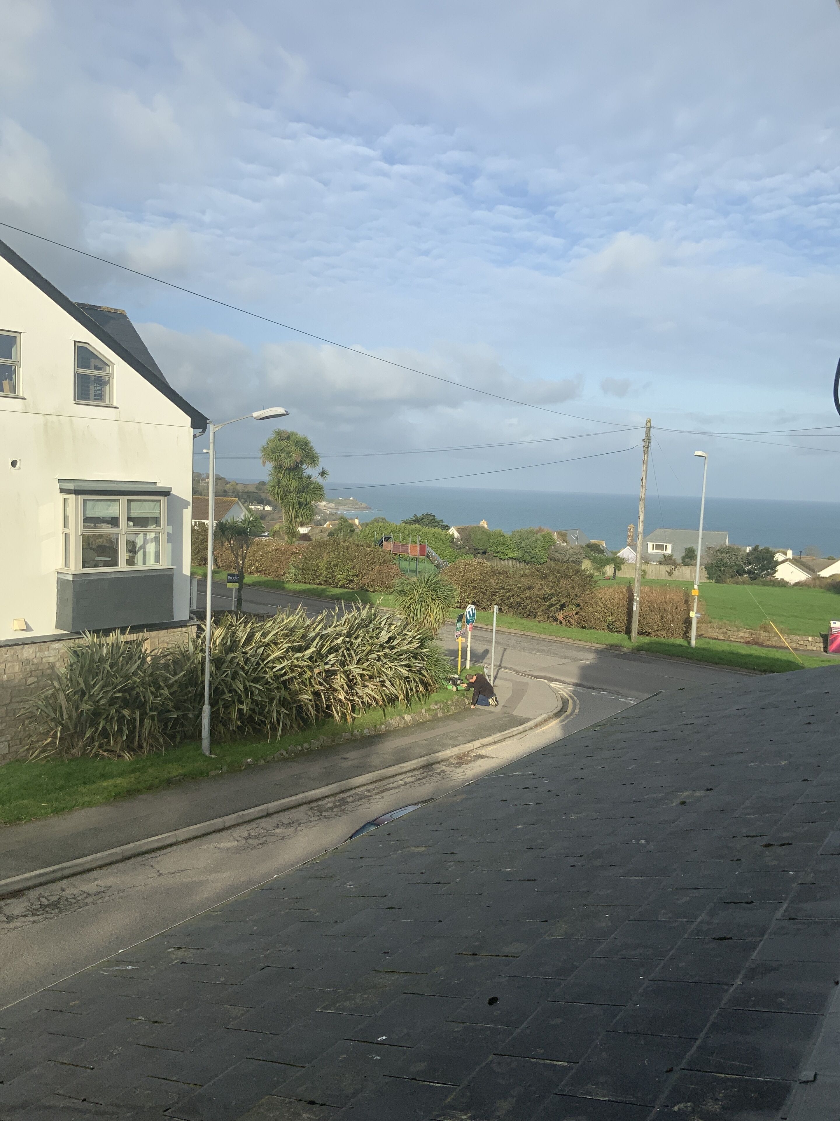 A residential street with a white house, a road curving to the right, and a view of the sea in the distance.