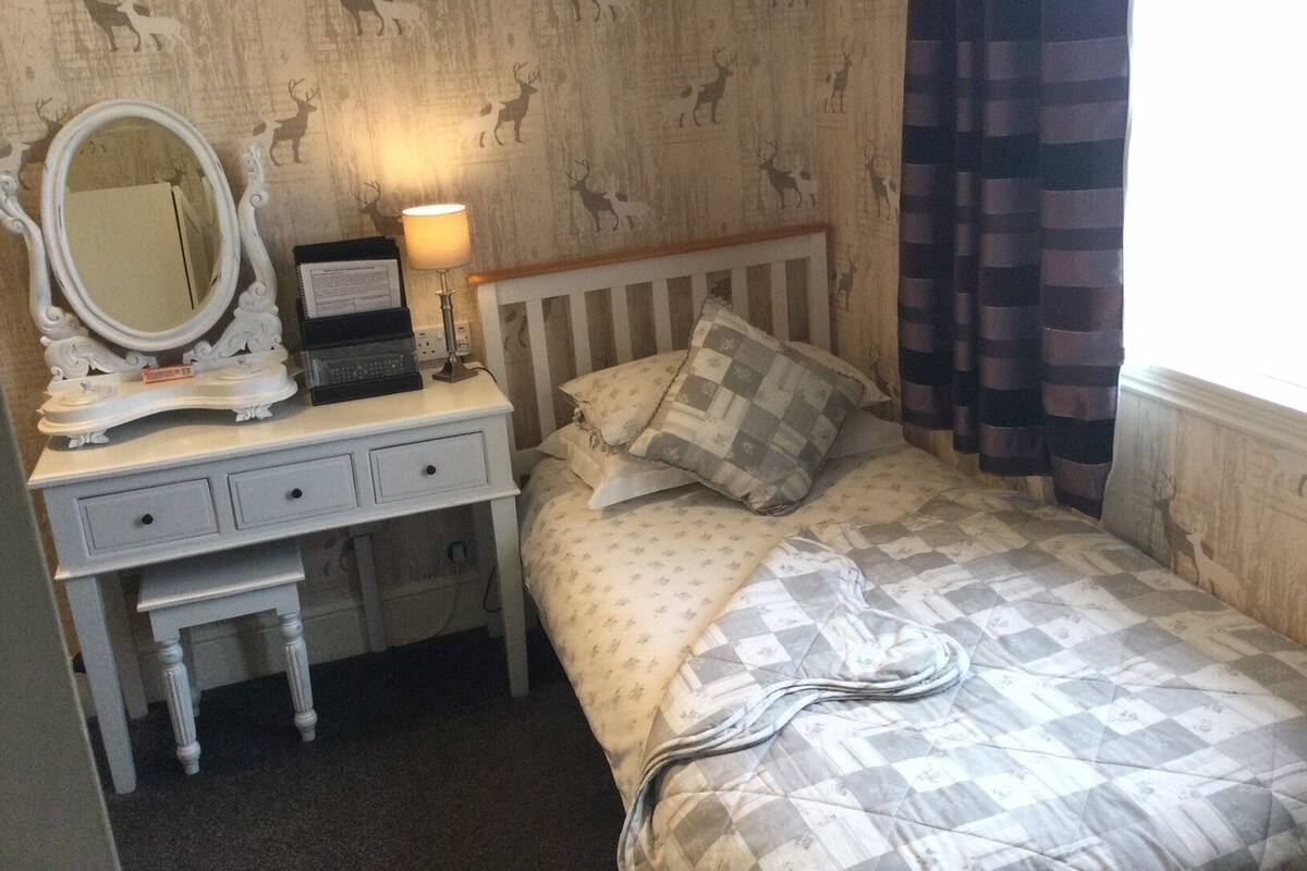 A bedroom with a bed, a nightstand with a lamp, a dresser with a mirror, and a patterned wallpaper.