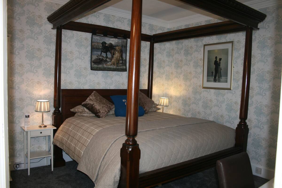 A four-poster bed with a quilted bedspread, two bedside tables with lamps, and framed pictures on the walls.