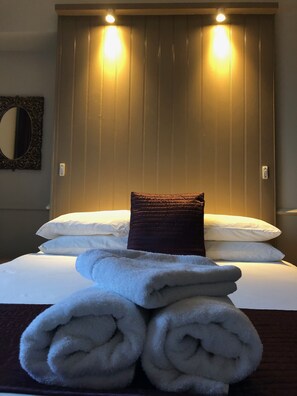 A neatly made bed with white linens and a dark brown pillow, a stack of white towels, and a mirror on the wall.
