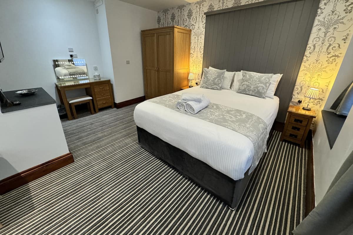 A hotel room with a large bed, a wooden wardrobe, a desk with a chair, and a small bedside table with a lamp.