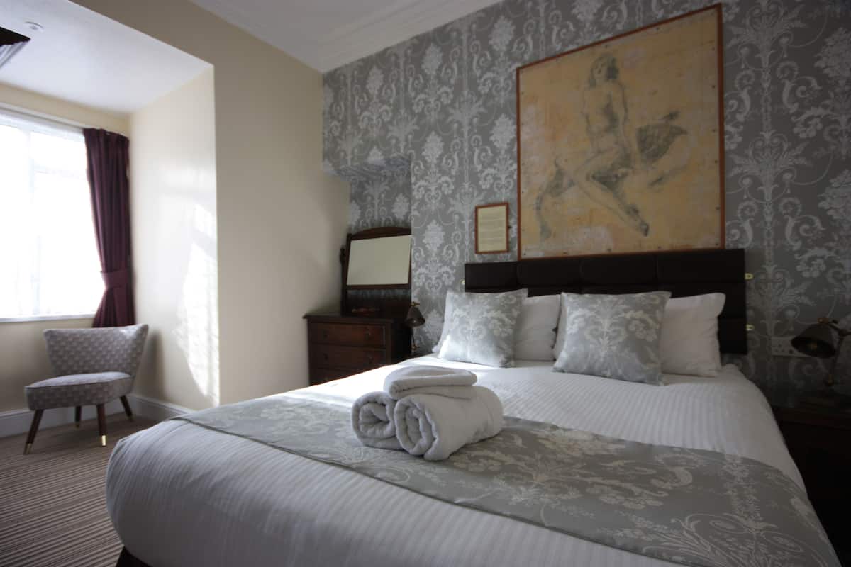 A neatly made bed with a rolled towel, a chair, a nightstand, and a painting on the wall.