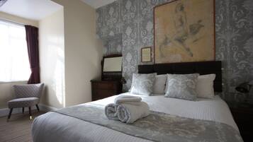 A neatly made bed with a rolled towel, a chair, a nightstand, and a painting on the wall.