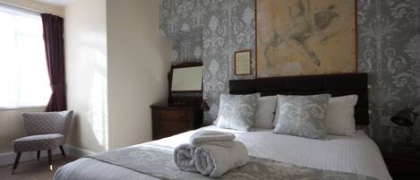 Una cama bien hecha, con una toalla enrollada, una silla, una mesita de noche y un cuadro en la pared.