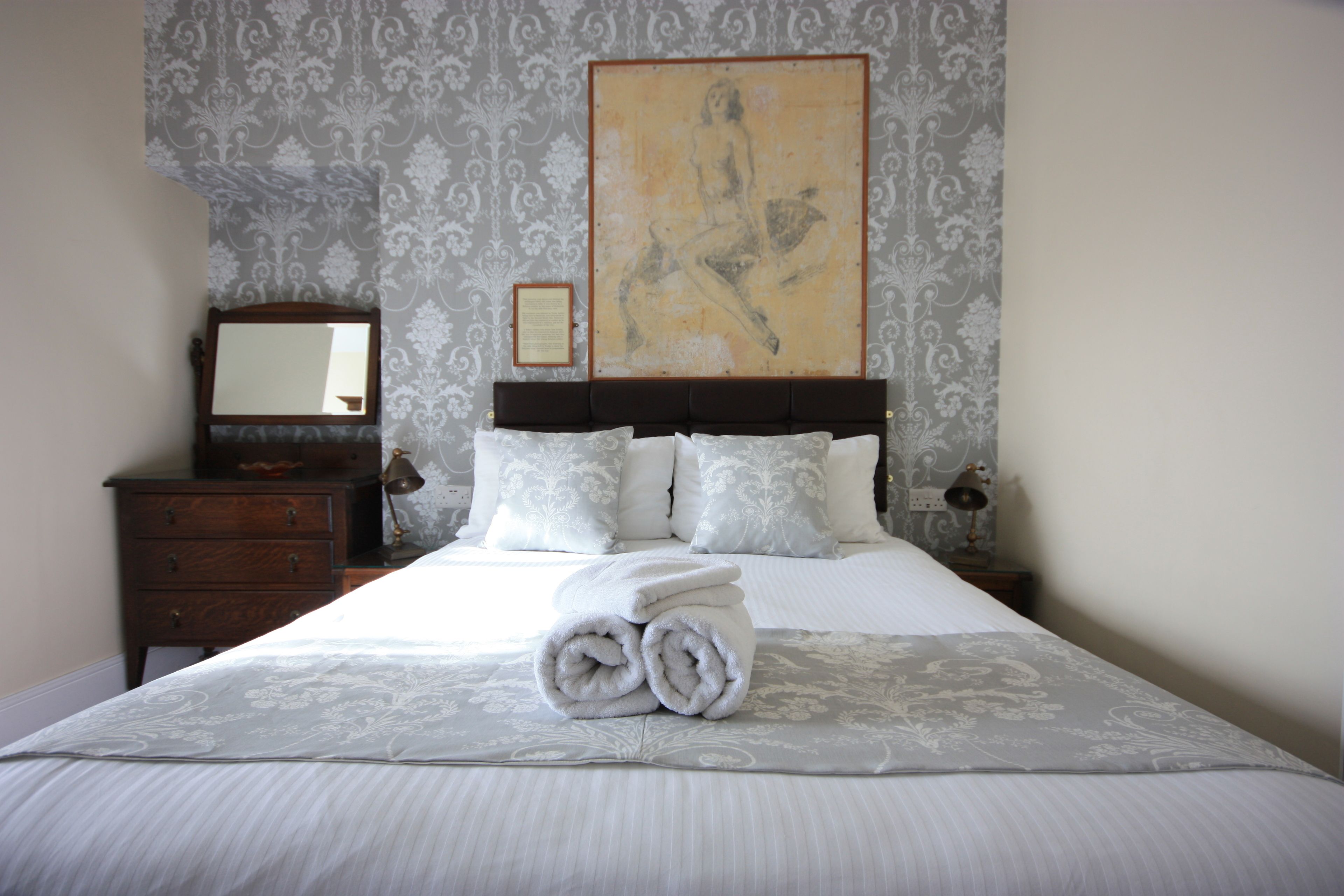 Una cama bien hecha con toallas enrolladas, una mesita de noche de madera con una lámpara, un aparador y una obra de arte grande enmarcada en la pared.