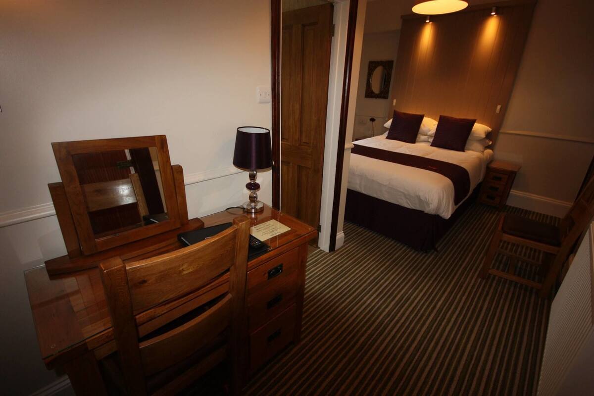 A hotel room with a wooden desk, a mirror, a lamp, and a bed with a headboard.