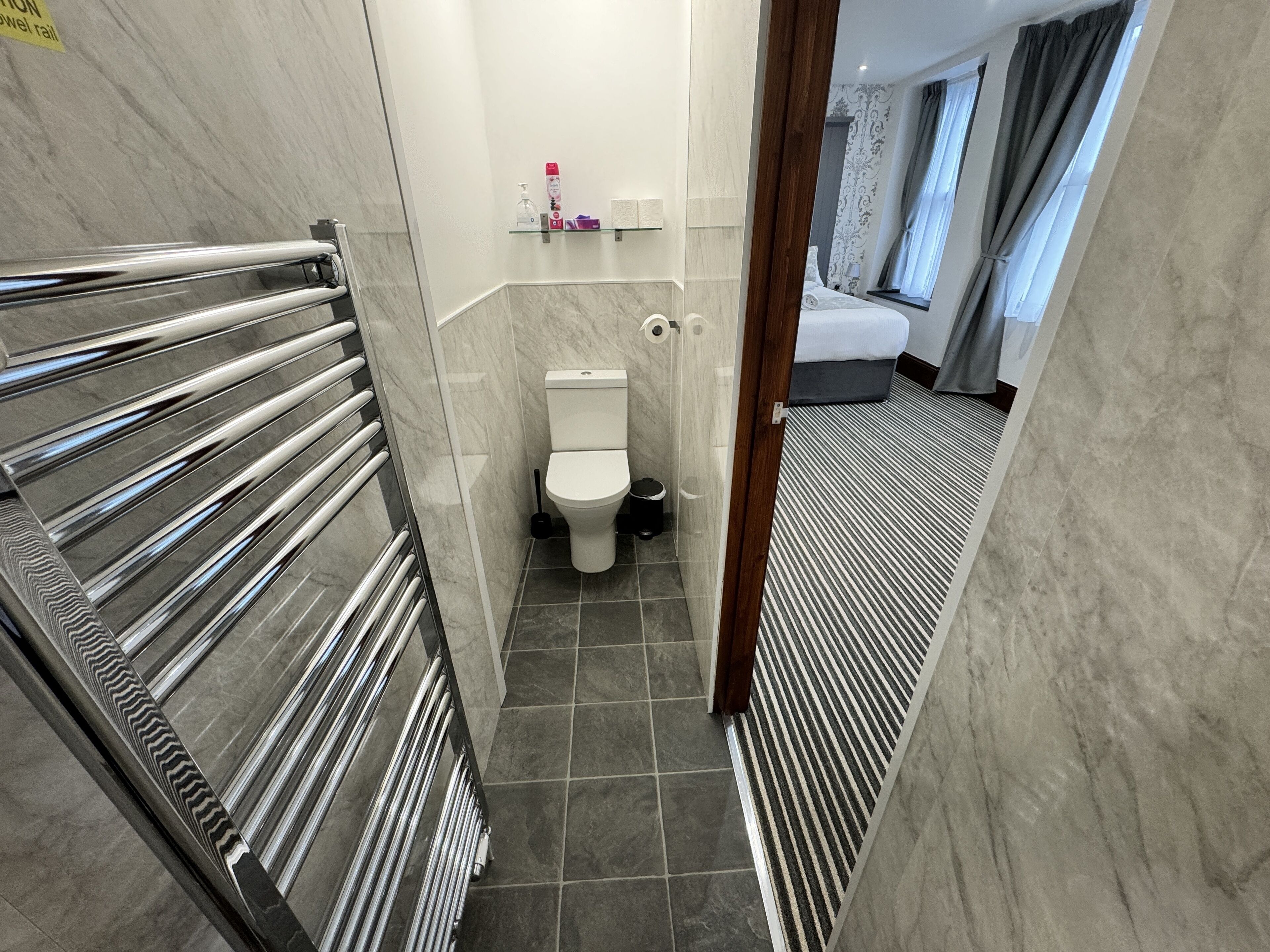 Un couloir étroit mène à une salle de bain équipée de toilettes, d’un sèche-mains et d’un espace douche.