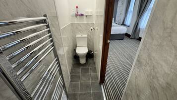 Un couloir étroit mène à une salle de bain équipée de toilettes, d’un sèche-mains et d’un espace douche.