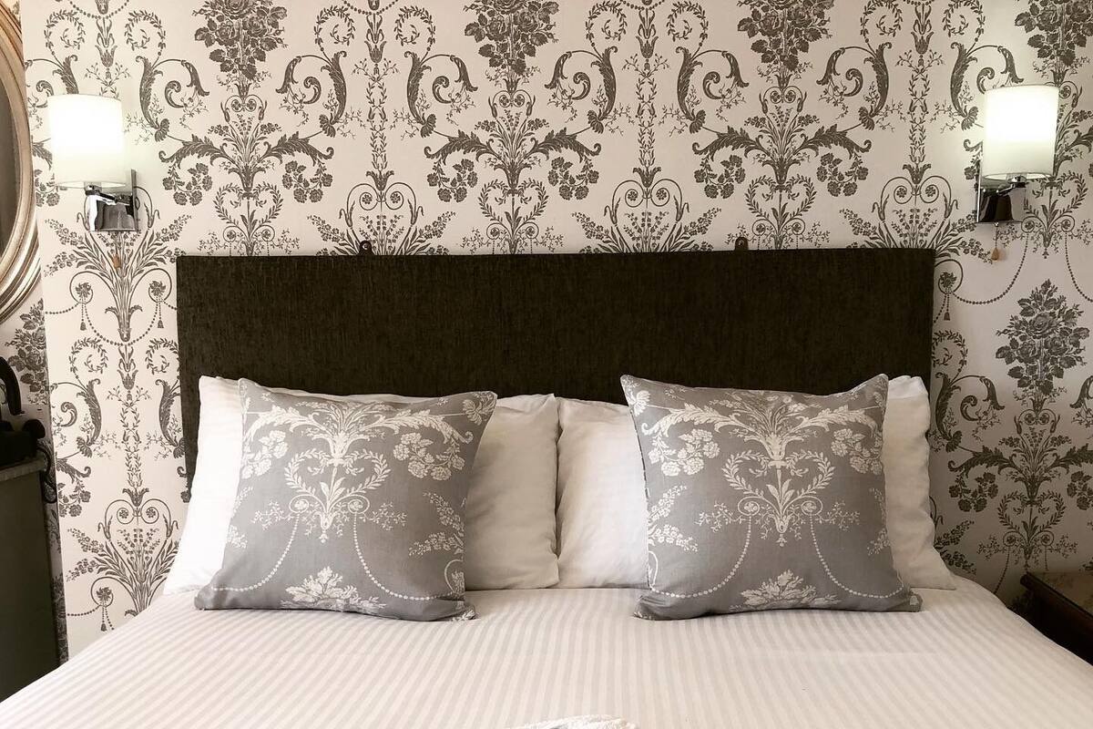A neatly made bed with two pillows and a folded towel, set against a wallpaper with a floral pattern.