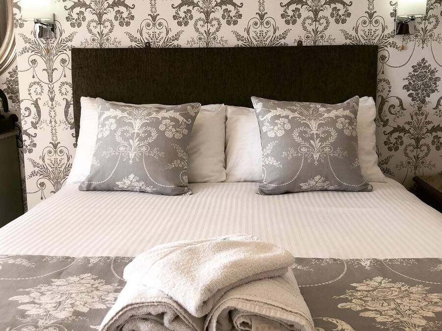 A neatly made bed with two pillows and a folded towel, set against a wallpaper with a floral pattern.