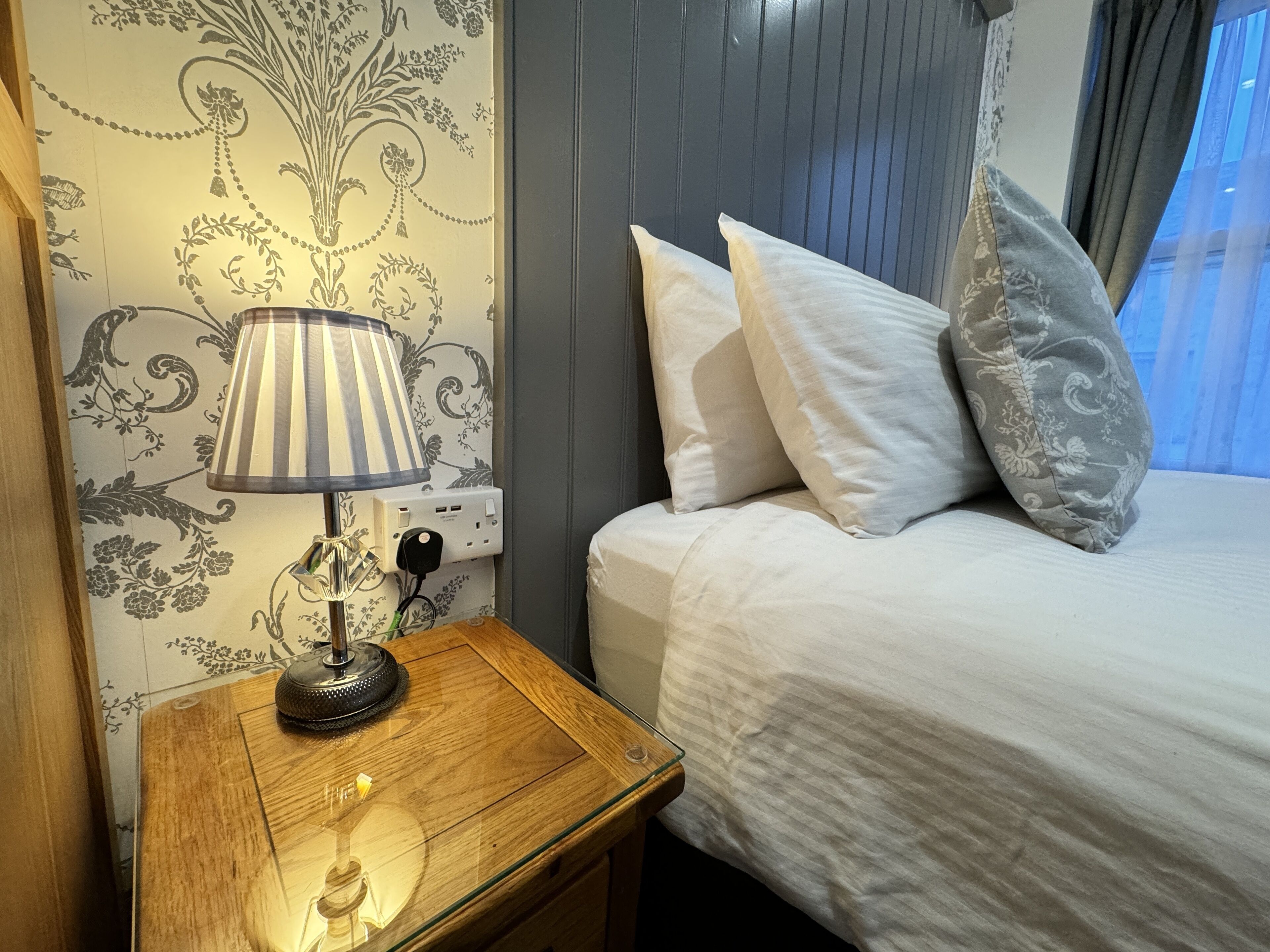 Un lit bien fait, avec des oreillers blancs et gris, une table de chevet en bois surmontée d’une lampe, et un mur orné d’un motif floral.