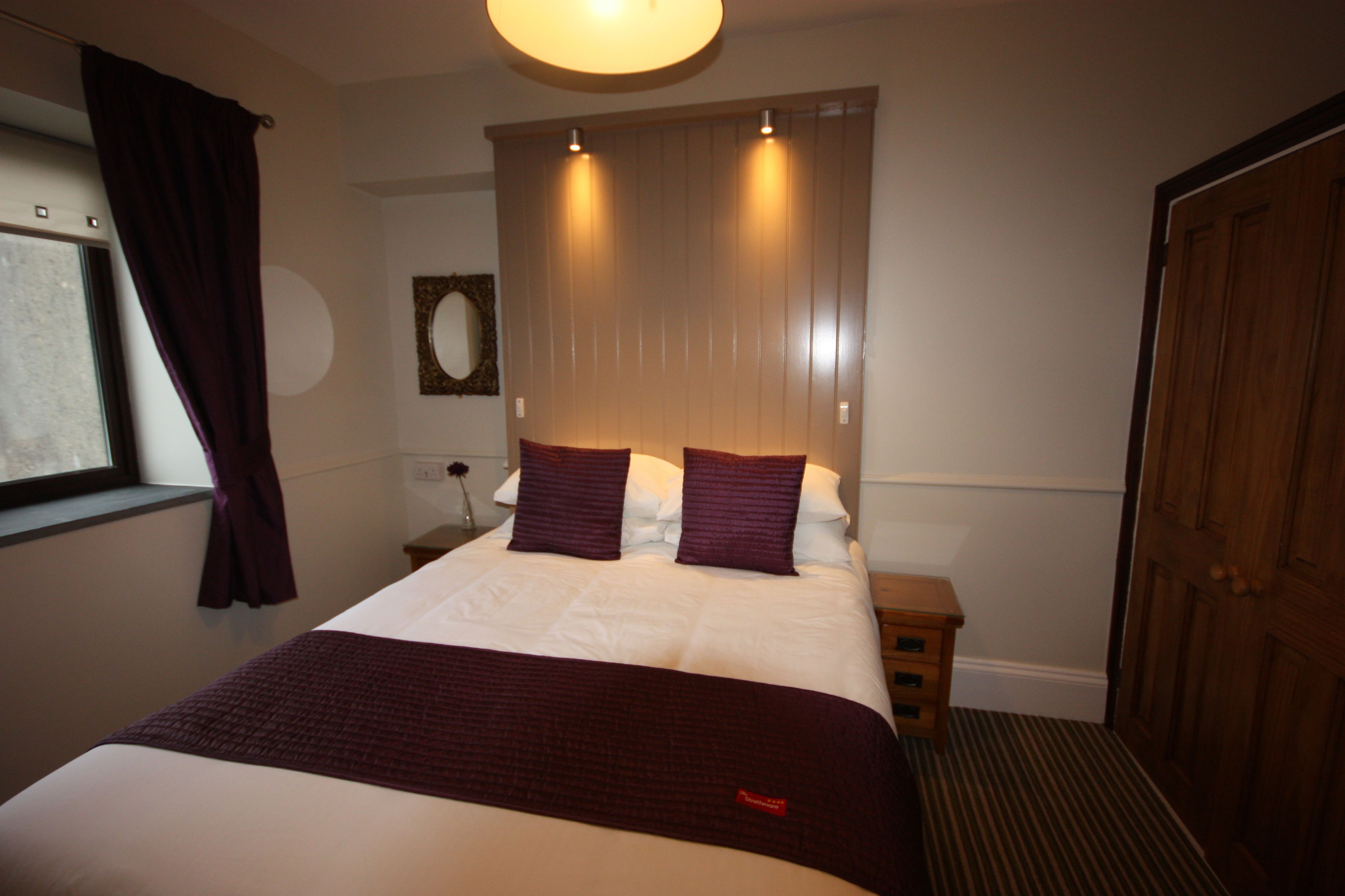 A neatly made bed with a white comforter and a dark red blanket, a wooden headboard with two lights, a wooden nightstand with a vase, a wooden door, and a window with purple curtains.