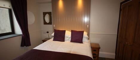 A neatly made bed with a white comforter and a dark red blanket, a wooden headboard with two lights, a wooden nightstand with a vase, a wooden door, and a window with purple curtains.