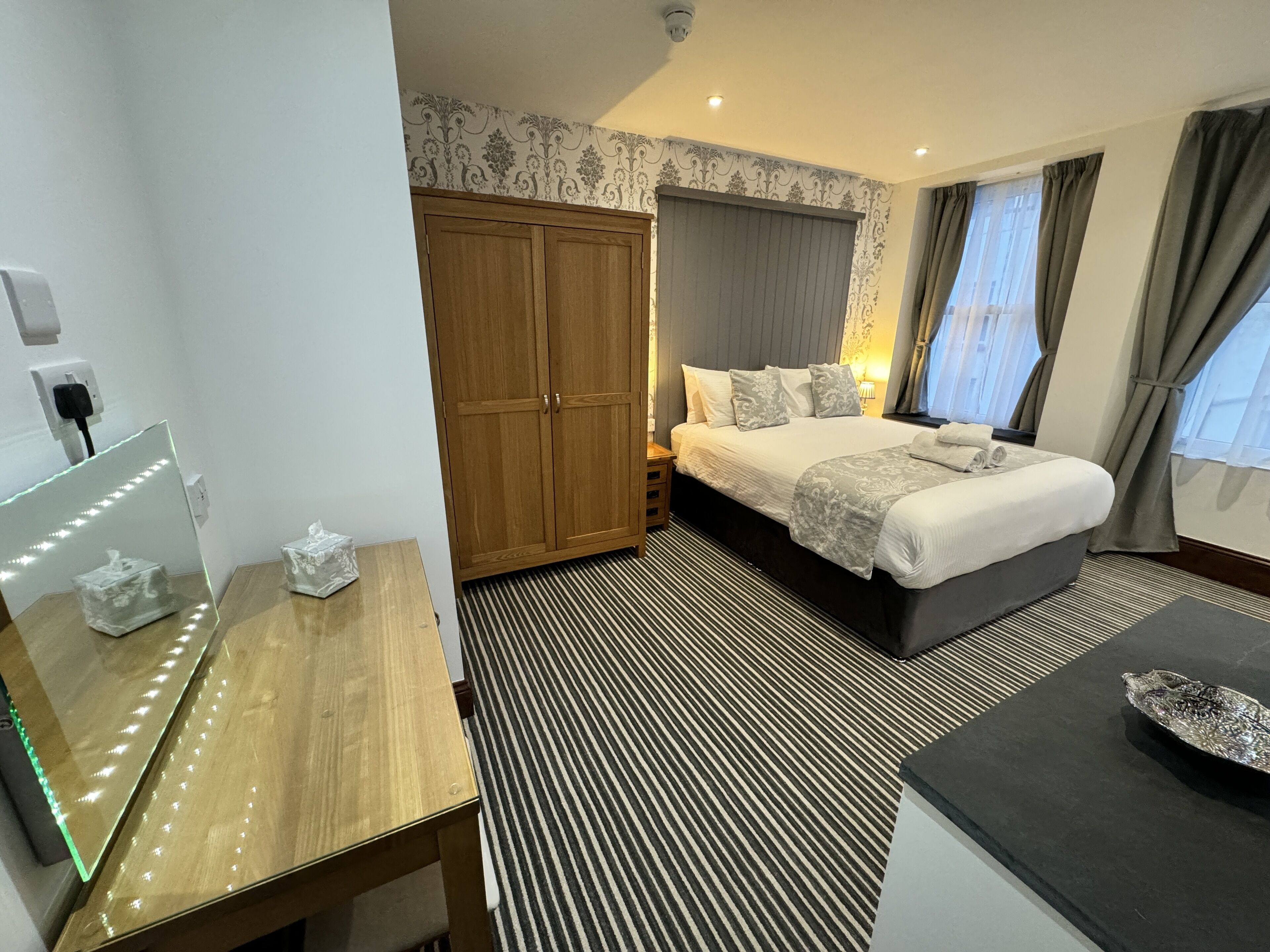 A hotel room with a large bed, a wooden wardrobe, a bedside table with a tissue box, a mirror, and a black and white striped carpet.