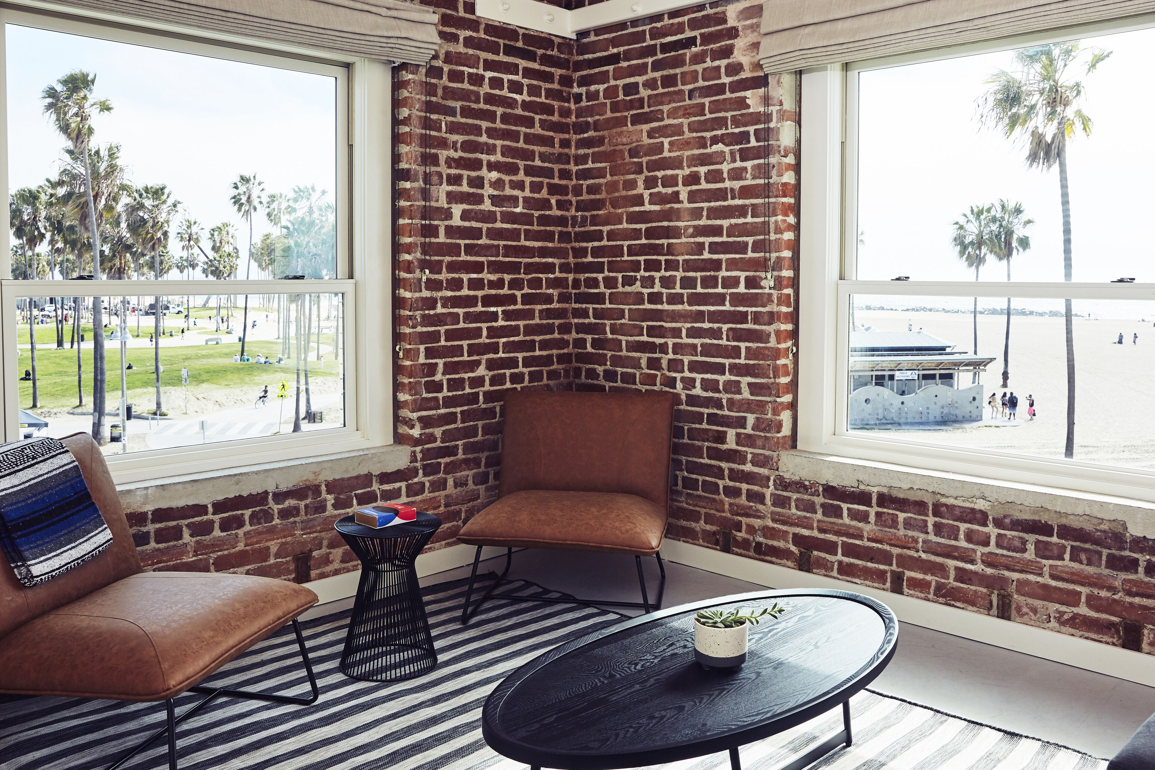 Habitación de esquina con pared de ladrillo, un sillón de cuero marrón, una mesa redonda negra y vistas a la playa.