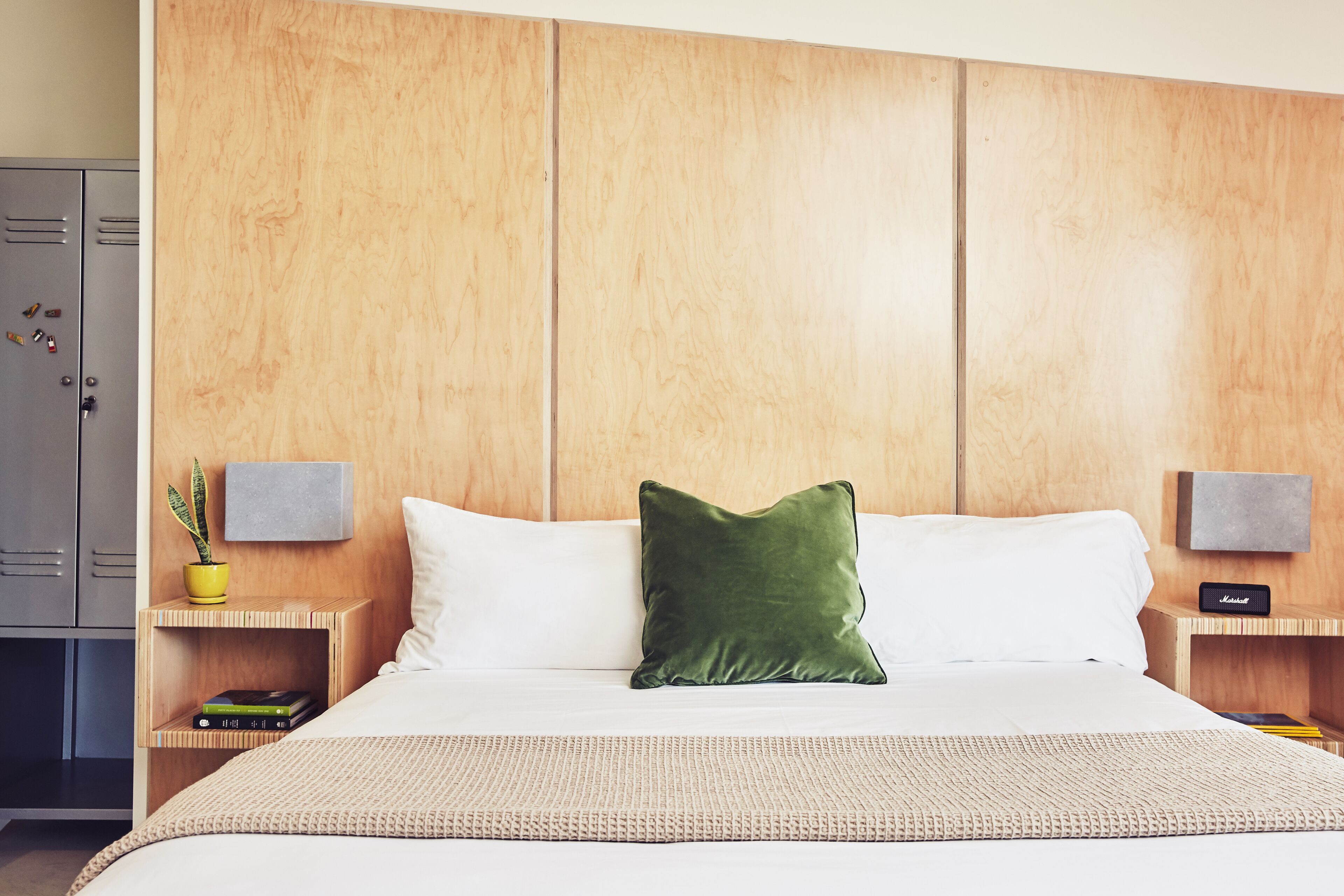 Une chambre d’hôtel avec un grand lit, deux tables de chevet, une tête de lit en bois, un oreiller vert, une petite plante et un haut-parleur Marshall.