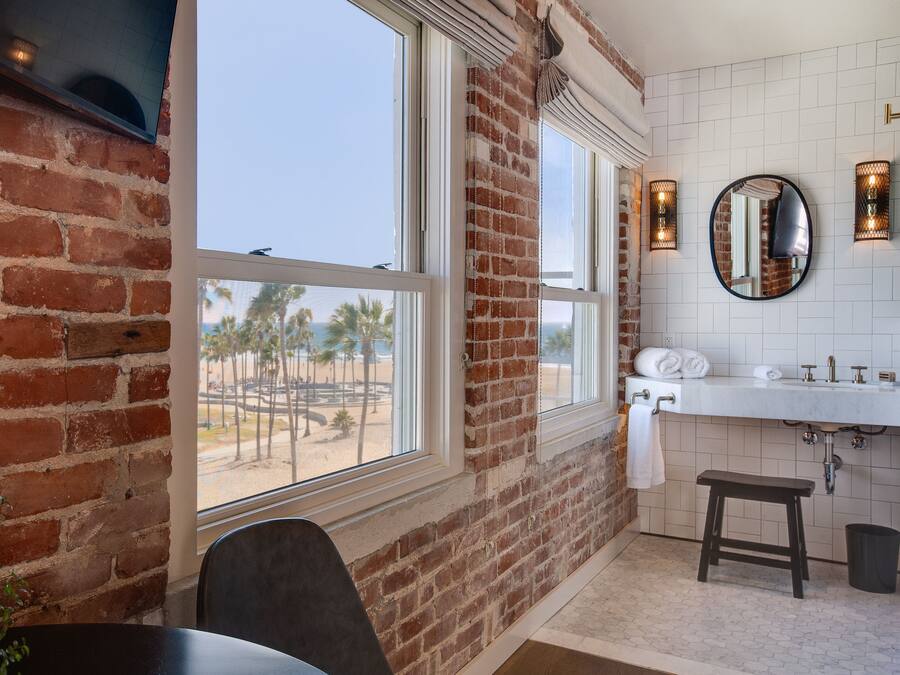 A bathroom with a brick wall, a large window with a view of palm trees, a white sink, and a round mirror.