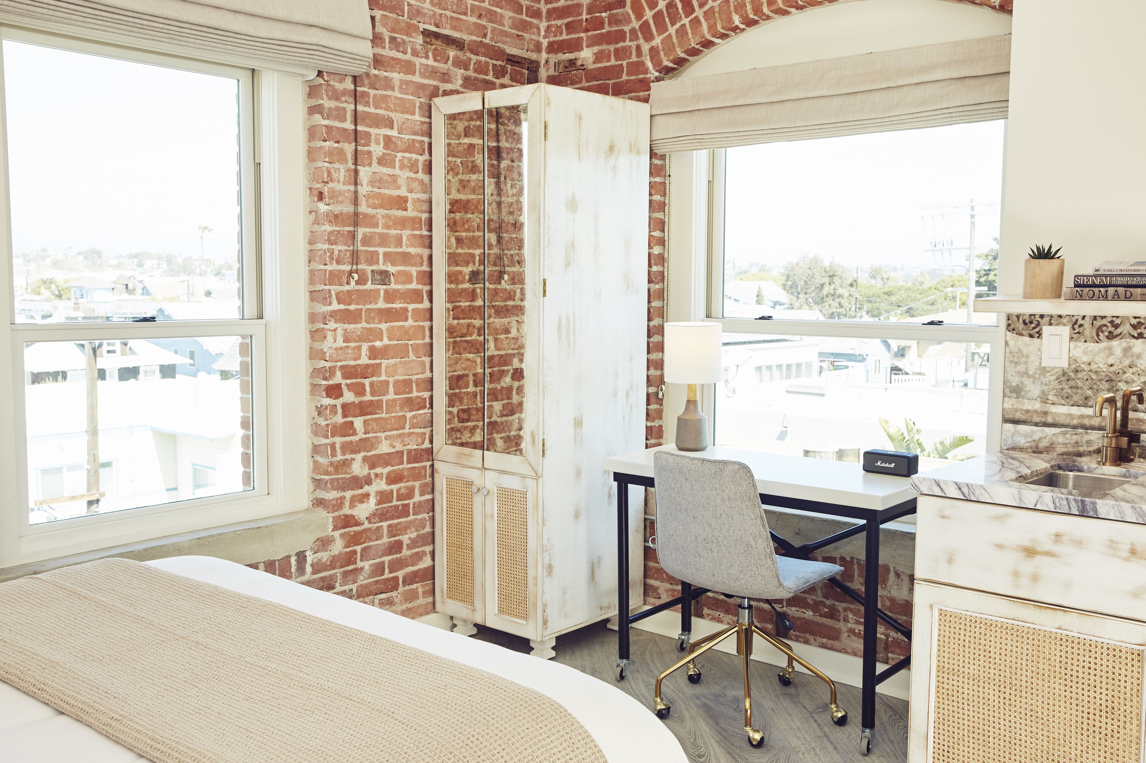 Een hotelkamer met een bakstenen muur, een witte kledingkast, een bureau met stoel, een bed en uitzicht op de stad door grote ramen.