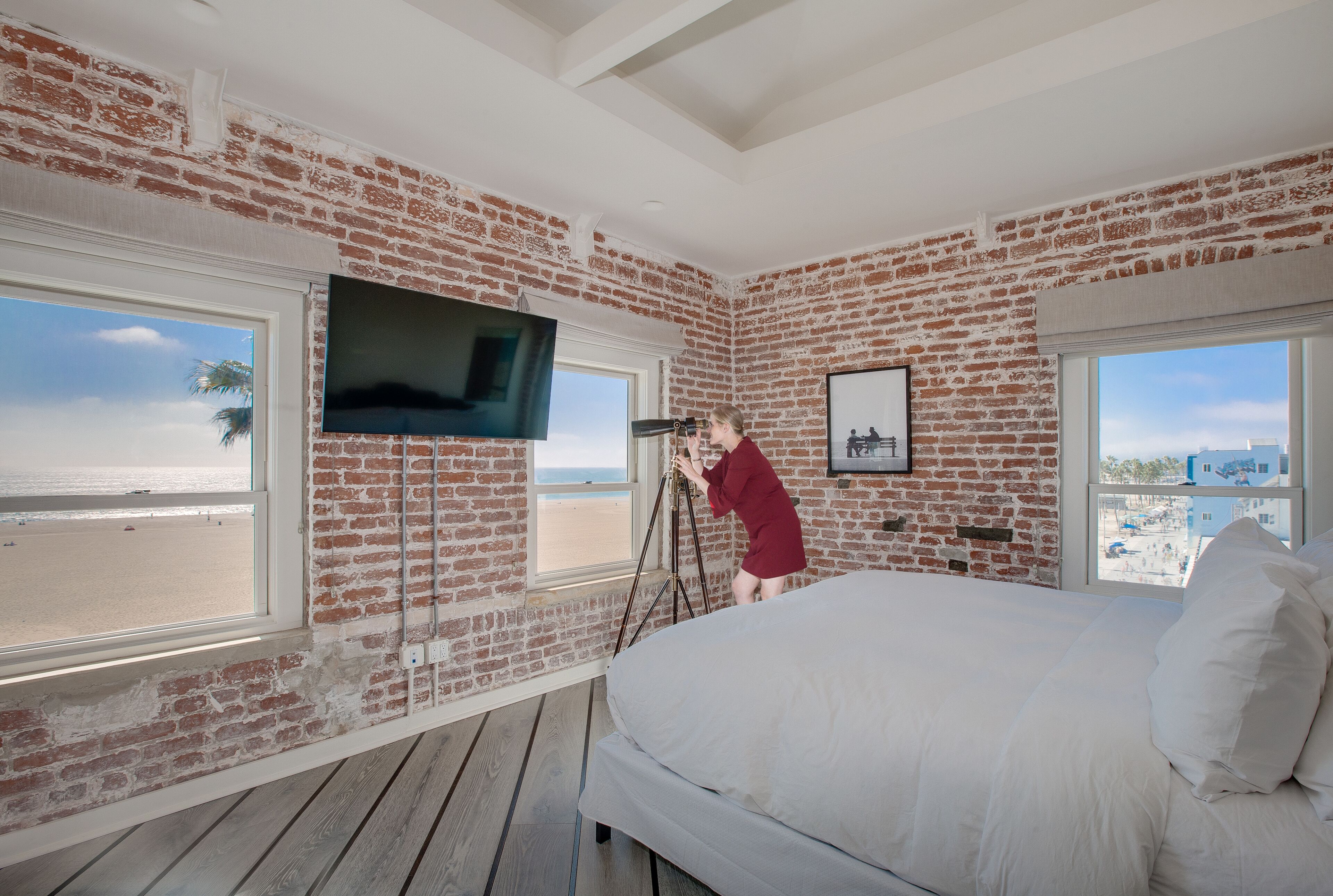 Un dormitorio con una cama grande, un televisor montado en la pared de ladrillo y una ventana con vistas a la playa.