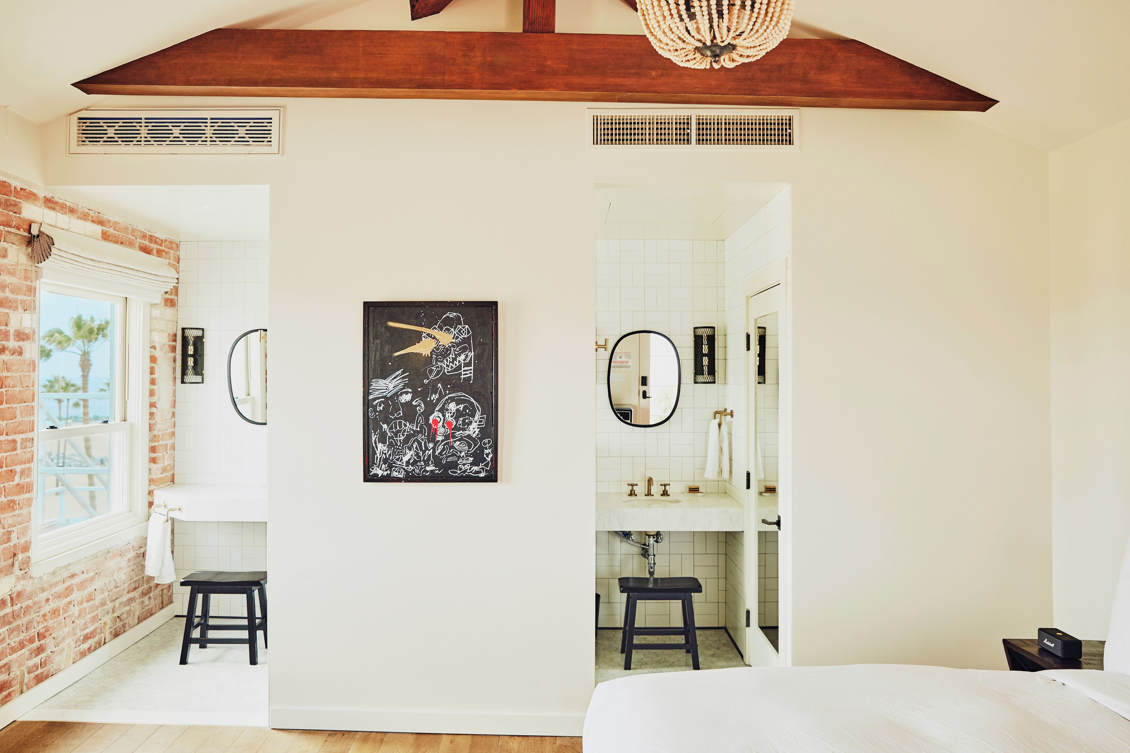 Una camera da letto con un letto grande, una finestra vista palme, un bagno con lavandino e specchio bianchi, e un'opera d'arte nera appesa al muro.