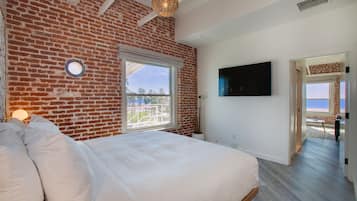 A bedroom with a brick wall, a large bed, a TV, and a view of the beach.