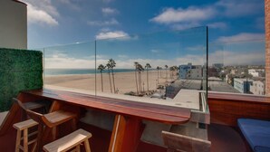 A balcony with a glass railing overlooking a beach, wooden bar stools, and a view of palm trees and buildings. - Venice V Hotel (Venice)