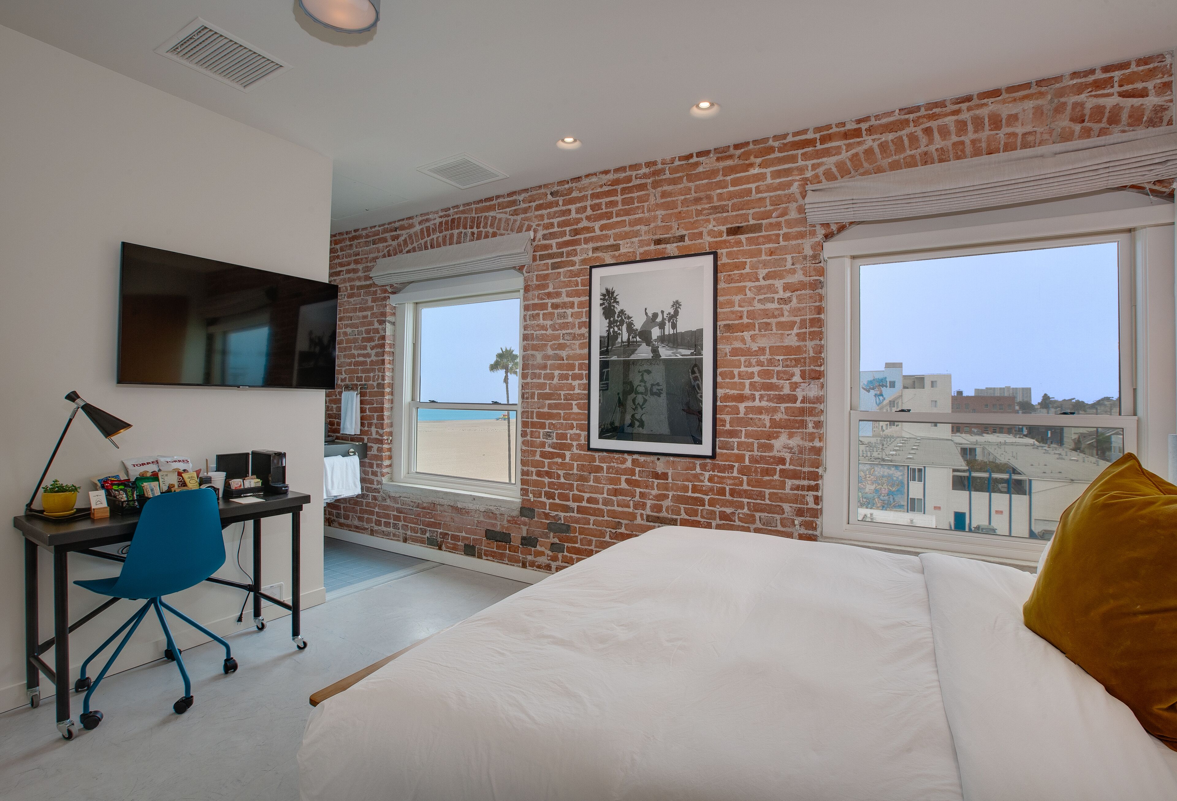 Une chambre avec un mur en briques, un lit, un bureau avec une chaise, une télévision et une fenêtre donnant sur la ville.