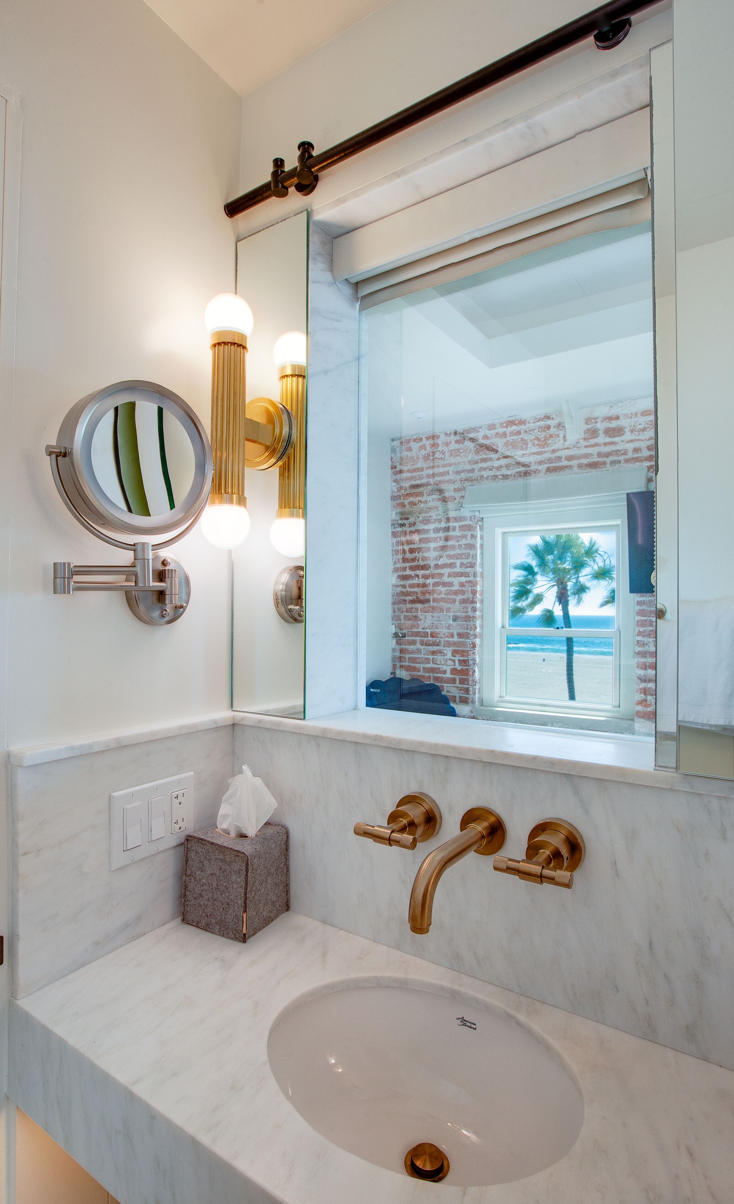 A bathroom with a marble sink, a round mirror, and a view of a brick wall and palm tree through a window.