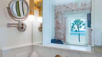 Une salle de bain avec un lavabo en marbre, un miroir rond et une vue sur un mur de briques et un palmier, le tout visible par une fenêtre.