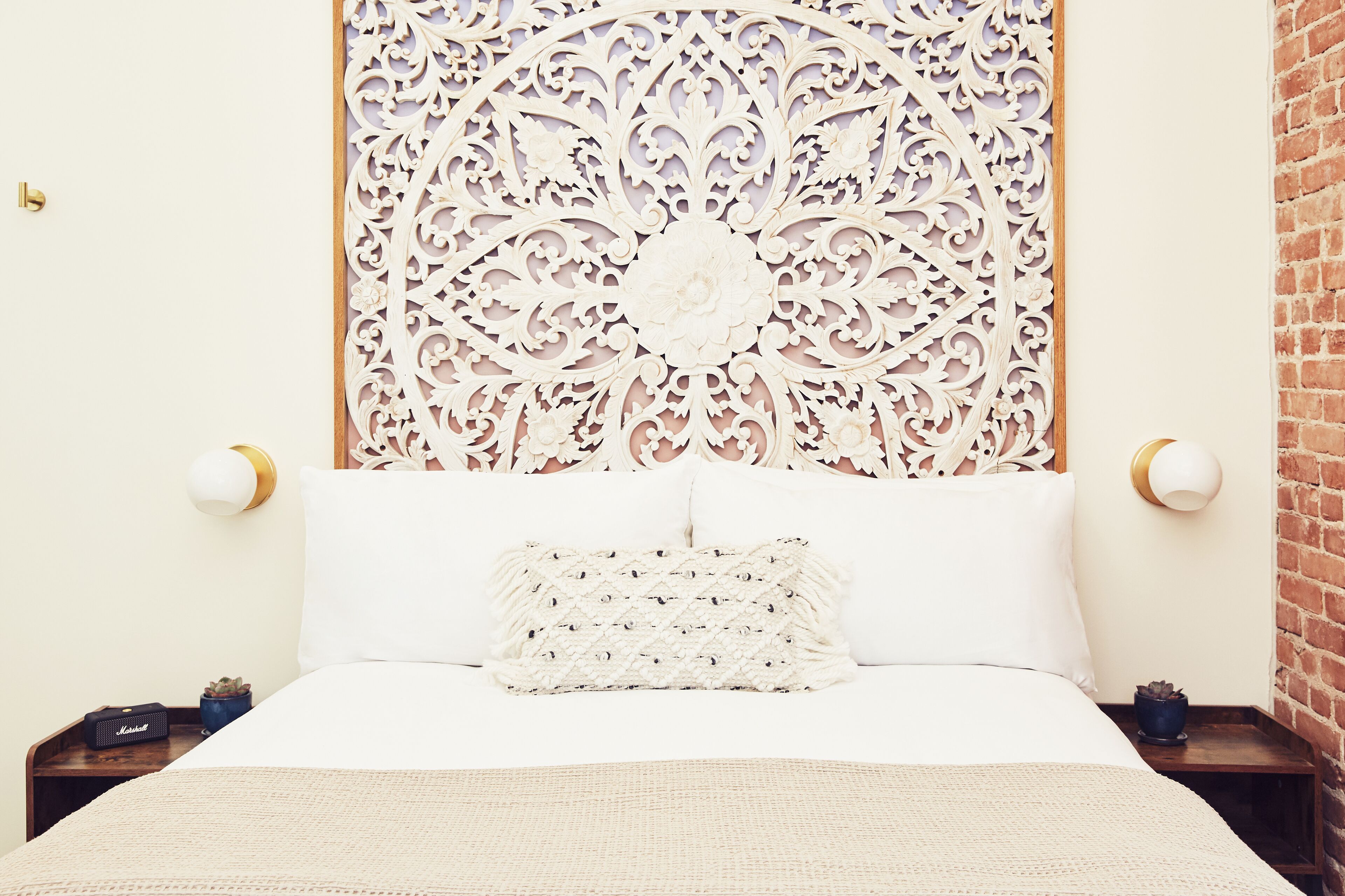 A neatly arranged bedroom with a decorative headboard, a white bed, and a small side table with a lamp and a plant.