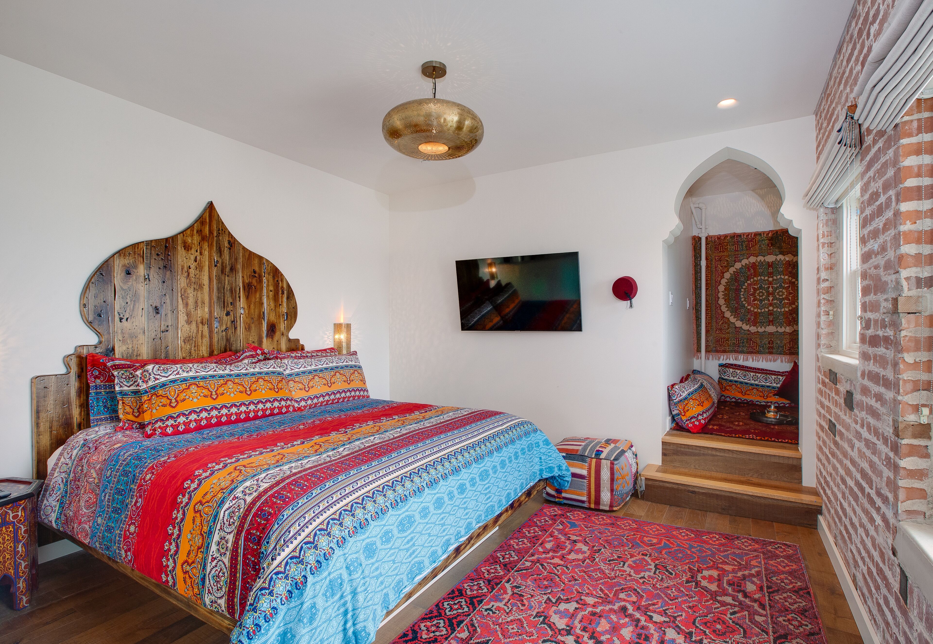A bedroom with a large bed, a wooden headboard, a red and blue patterned bedspread, a red rug, a small table with a lamp, and a TV on the wall.