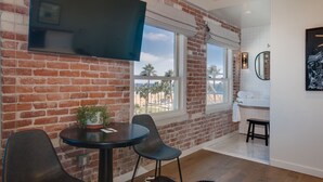 A room with brick walls, a large window, a flat-screen TV, a round table, two chairs, a small plant, a black and white rug, a white bathtub, and a mirror. - Venice V Hotel (Venice)