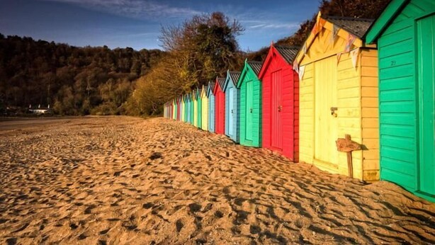 A row of colorful beach huts along a sandy beach with a forested hill in the background. - Craig Y Glyn (Pwllheli)