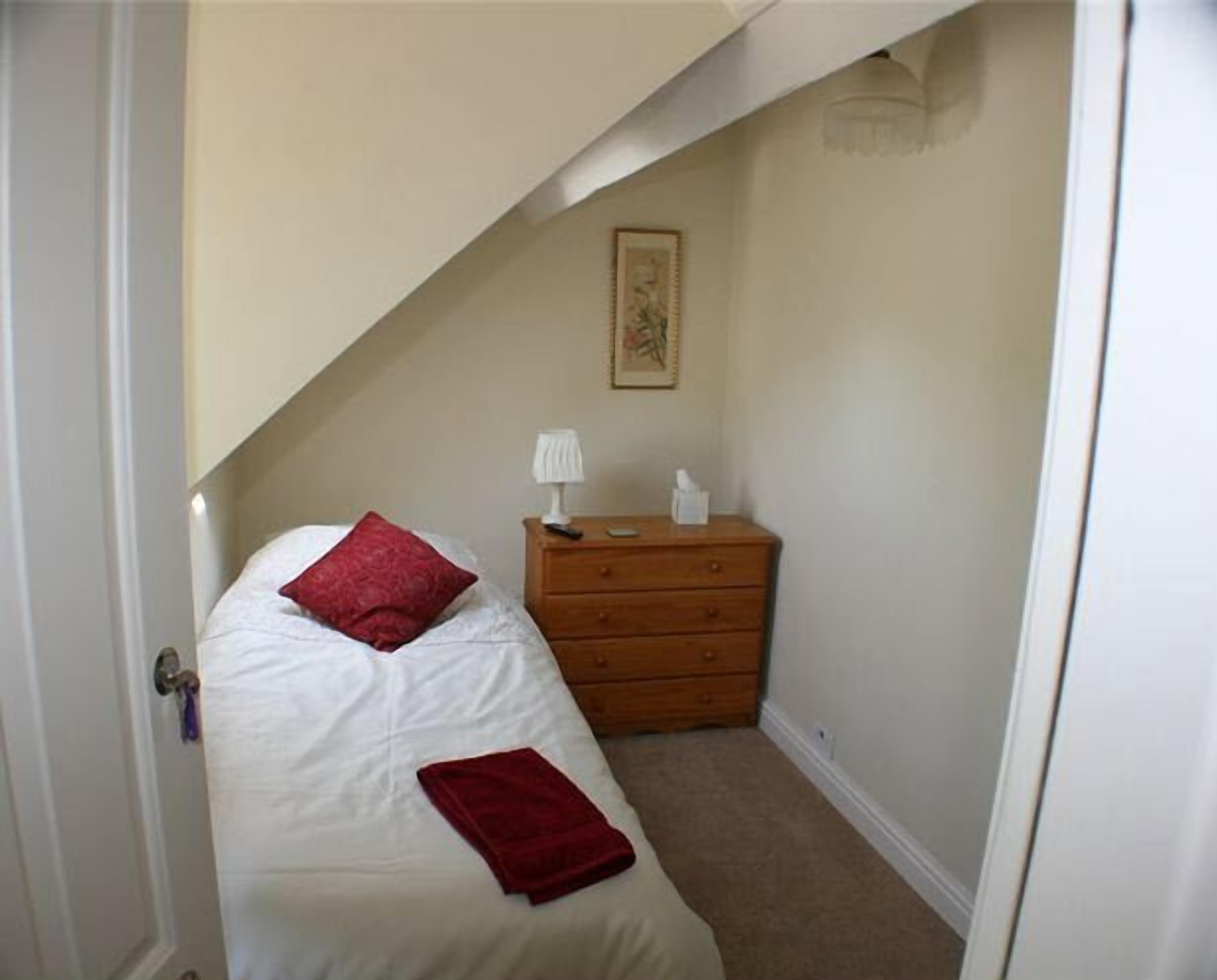 A small, narrow room with a single bed, a wooden dresser, and a framed picture on the wall.