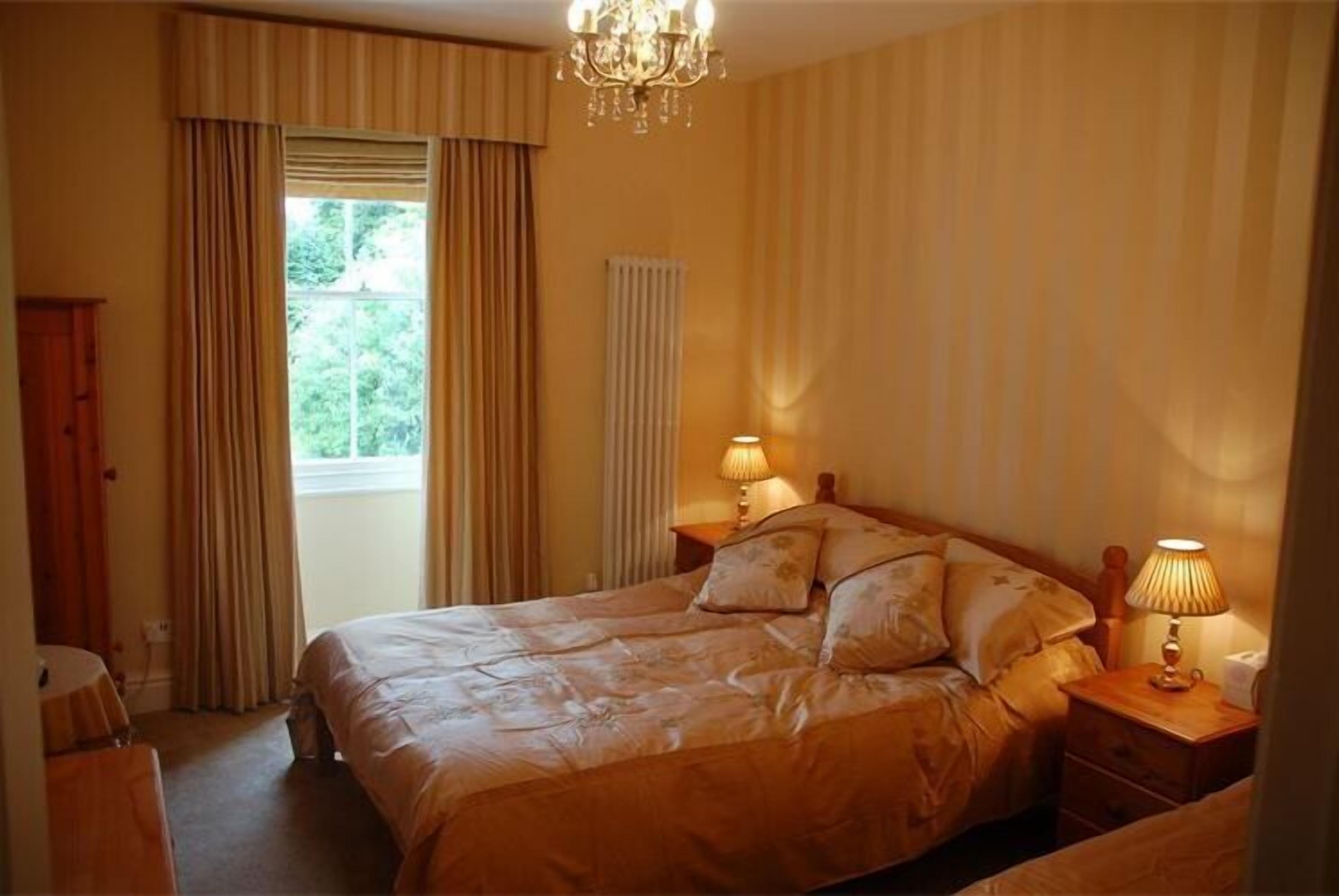 A bedroom with a large bed, two bedside tables with lamps, a window with curtains, and a radiator.