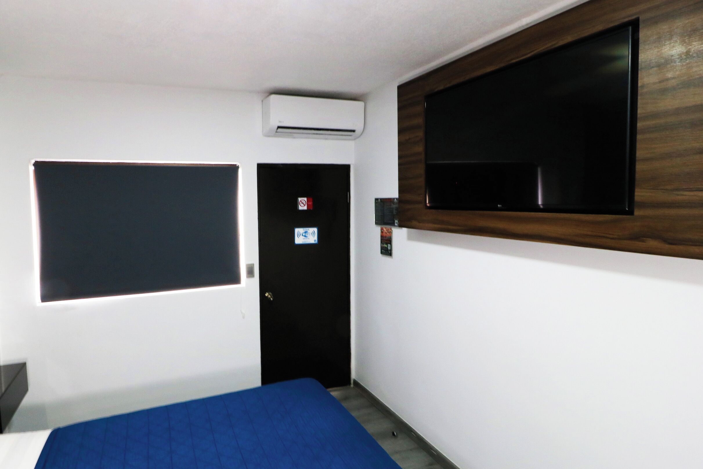 Habitación de hotel con cama, televisor de pantalla plana en la pared, equipo de aire acondicionado y puerta con letrero de no fumar.