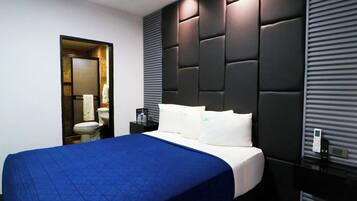 Una habitación de hotel moderna con una cama grande, una mesita de noche, un baño visible a través de una puerta abierta y un panel de control en la pared.