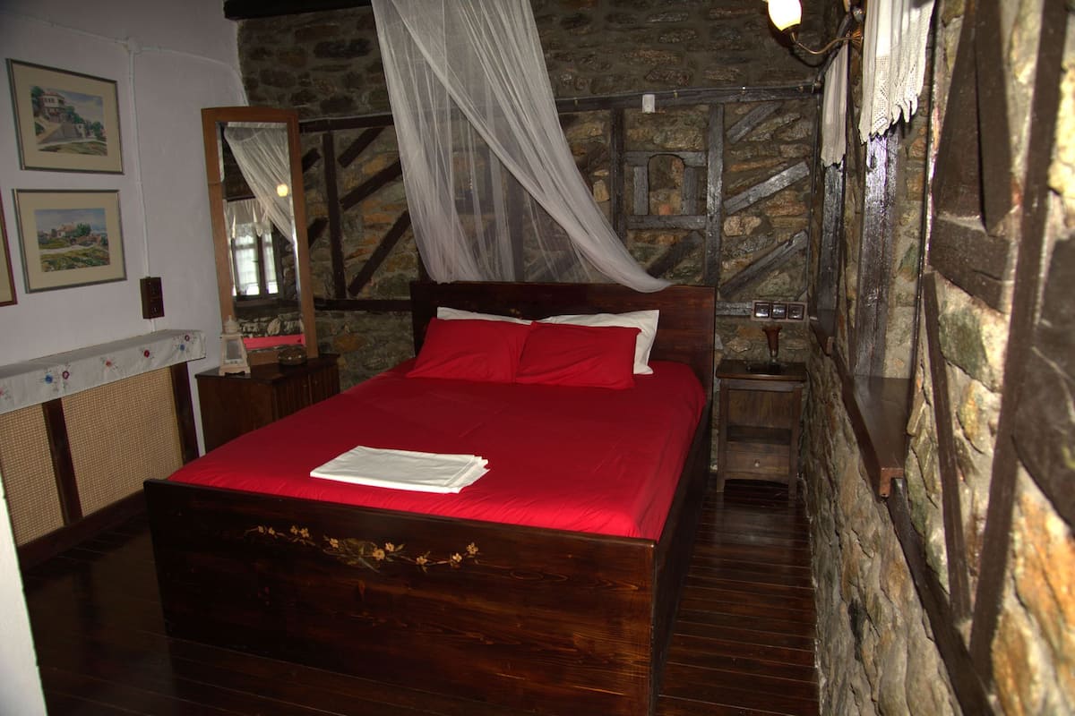 Una cama de madera con colcha roja y almohadas blancas, cabecera de madera y pared de piedra.